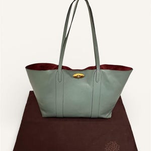 May include: A light blue leather tote bag with long handles and a gold-colored clasp. The interior is a deep burgundy color. The bag is sitting on a brown fabric dust bag with a tree logo.