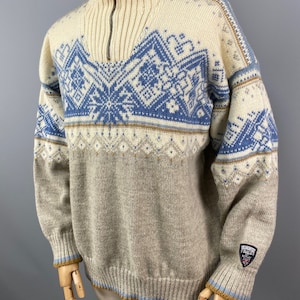 May include: A beige and blue patterned sweater with a half-zip closure. The sweater has a brown and blue trim around the neck and cuffs. The sweater has a small patch on the lower right side with the text "Genuine Italy" in white.