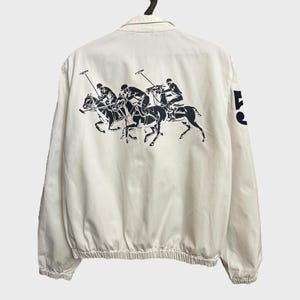 May include: A white long-sleeve jacket with a navy blue polo player graphic on the back. The jacket has a zipper closure and a drawstring at the bottom.