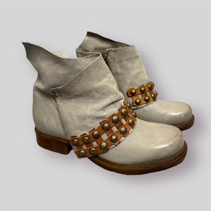 May include: A pair of gray ankle boots with brown leather straps and gold studs. The boots have a slouchy, casual look.