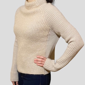 May include: A beige knitted sweater with a mock neck and long sleeves. The sweater has a ribbed texture and a slightly fitted silhouette.