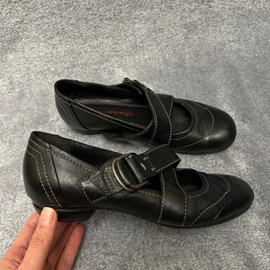 Może przedstawiać: Para czarnych sk&oacute;rzanych but&oacute;w z klamrą i małym obcasem. Buty mają białe przeszycia i widoczne jest słowo "Step" wewnątrz jednego buta.