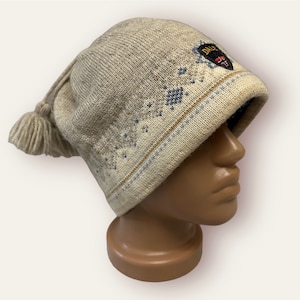 May include: A beige knit hat with a tassel and a patterned design. The hat features a blue and brown geometric pattern and a patch with the word "DALE" and a crest. The hat is displayed on a mannequin head.