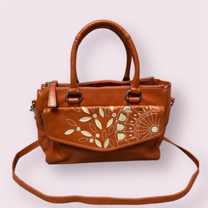 May include: A brown leather satchel handbag with a white floral design. The bag has a top handle and a long shoulder strap.