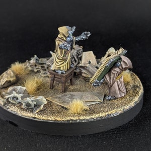 May include: Two miniature figures in hooded robes stand on a circular base covered in sand and debris. One figure is wearing a brown robe and holding a book, while the other figure is wearing a yellow robe and holding a staff. The base is decorated with small rocks and tufts of grass.