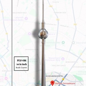 May include: A silver-coloured metal model of the Berlin TV Tower featuring a spherical observation deck and a tall antenna spire. The model measures 37.5 cm in height with a 1:1000 scale, displayed against a map of Berlin, Germany.