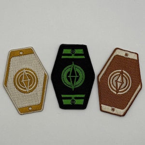 Puede incluir: Tres parches bordados en forma hexagonal. Uno es beige con detalles dorados, otro es negro con detalles verdes y el tercero es marrón con detalles dorados. Cada uno presenta un diseño geométrico central.