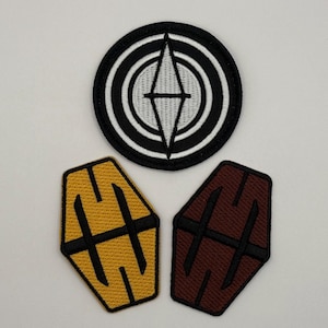 May include: Three embroidered patches. One round patch with a black and white concentric circle design and a diamond shape in the center. Two hexagonal patches, one gold with a black design, and one maroon with a black design.