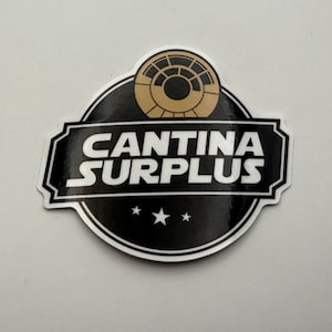 May include: A black and white sticker featuring the words "CANTINA SURPLUS" in white, with three white stars at the bottom. The top of the sticker has a gold and black circular design. The sticker has a white border.