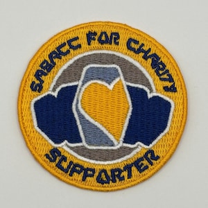Pode incluir: Um remendo circular bordado amarelo com o texto "Sabacc for Charity Supporter" em um círculo branco com um coração azul e cinza dentro.