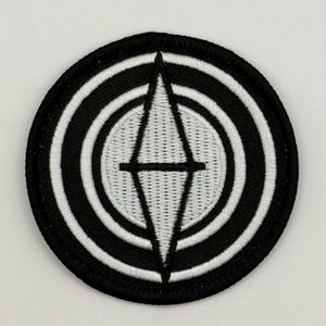 May include: Black and white embroidered patch with a diamond shape in the center surrounded by concentric circles.