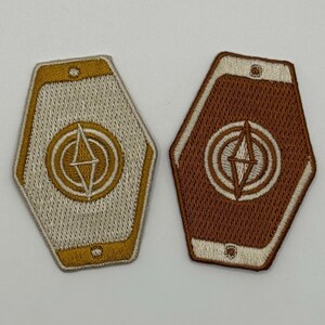 May include: Two embroidered patches with a geometric design. One patch is beige with gold accents, and the other is brown with cream accents. Both have a central compass-like emblem within concentric circles.