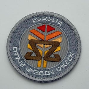 May include: Circular, grey embroidered patch with a central design. The design features a brown and yellow symbol, topped by a red, orange, and yellow triangle. The patch has text in a circular pattern around the design.