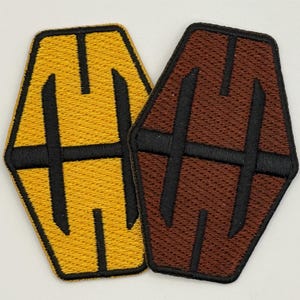 May include: Two embroidered patches in the shape of a coffin. One patch is yellow with black stitching and the other is brown with black stitching.