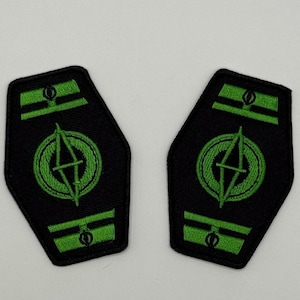 以下が含まれることがあります： 緑色の円形デザインと稲妻が施された、2つの黒い刺繍パッチ。パッチには、上部と下部に緑色の水平バーがあります。パッチは白い背景にあります。