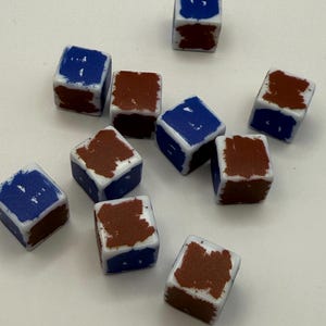 May include: Ten white dice with blue and brown paint splatter designs. The dice are all in focus and are arranged in a random pattern on a white surface.