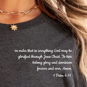 May include: Dark gray t-shirt with the text "-in order that in everything God may be glorified through Jesus Christ. To him belong glory and dominion forever and ever. Amen. 1 Peter 4:11." A gold necklace is visible.