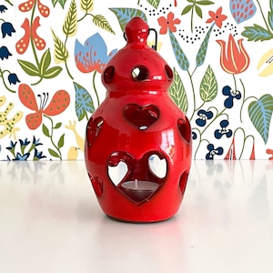 May include: A glossy red ceramic lantern with heart-shaped cutouts. The lantern has a cylindrical shape with a rounded top and a small handle. A lit tea light candle is visible inside. The background features a floral wallpaper.