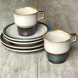 May include: Two ceramic coffee mugs with a brown and blue glaze. The mugs are stacked on top of each other. The mugs are sitting on a set of three saucers with a blue and brown trim.