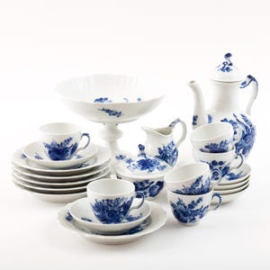 Op de afbeelding: Een compleet wit porseleinen theeservies met blauwe bloemmotieven. De set bevat kopjes, schotels, borden, een suikerpot, een melkkan, een serveerschaal en een theepot. De theeset staat op een wit oppervlak.