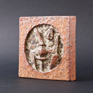 May include: A brown ceramic tile with a raised relief design of a person sitting in a chair. The tile has a speckled brown and white glaze.