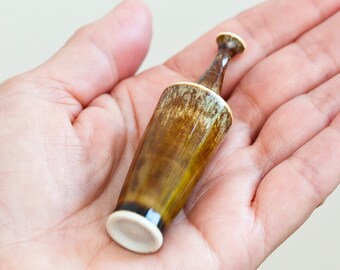 Puede incluir: Una botella de cerámica en miniatura con un esmalte brillante moteado en tonos marrón y verde oliva. El pequeño recipiente tiene un cuello cónico, un borde ensanchado y una base blanca plana sin esmaltar, con una superficie lisa.