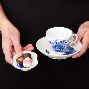 May include: A white teacup and saucer with a blue floral design. The saucer has a scalloped edge. Three small chocolate candies are on a small white plate.