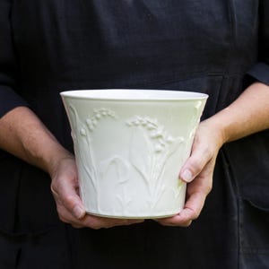 Puede incluir: Una maceta de cerámica blanca con un diseño en relieve de flores de lirio de los valles. La maceta está siendo sostenida por las manos de una persona.