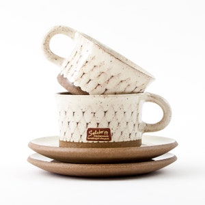 Puede incluir: Dos tazas de café y platillos de cerámica apilados. Las tazas y los platillos son de color blanquecino con un patrón texturizado y bordes marrones. Las tazas tienen asas. Una etiqueta con texto es visible en la taza delantera.