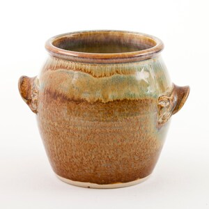 Puede incluir: Una olla de cerámica marrón con dos asas. La olla tiene un esmalte jaspeado con tonos marrones, verdes y beige.