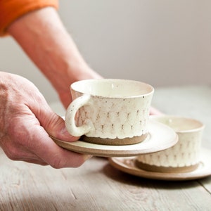 Puede incluir: Dos tazas de té de cerámica blanca con motas marrones y un borde ondulado. Las tazas están sobre platillos a juego.
