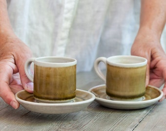 May include: Two ceramic cups and matching saucers featuring a cylindrical shape with a handle. The glaze is a gradient of earthy olive green and brown tones, transitioning to a lighter, off-white cream color at the top rim and the base.