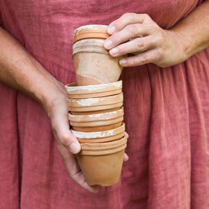 May include: A stack of six terracotta flower pots, with the top pot slightly tilted to the side. The pots are all different sizes and have a rustic, earthy look.