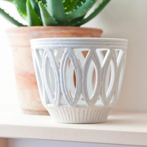 Puede incluir: Maceta de cerámica blanca con un diseño decorativo de corte. La maceta tiene una base ancha y redonda y una parte superior ligeramente acampanada. Los recortes tienen forma de óvalos y están dispuestos en un patrón repetitivo.