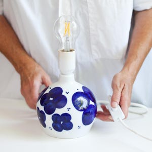 May include: A white ceramic table lamp with a round base, featuring a pattern of blue floral designs. The lamp has a white bulb socket and a clear light bulb. A white cord with an on/off switch is attached.