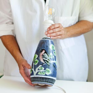 Può includere: Lampada da tavolo in ceramica blu con un disegno di uccello e fogliame. La lampada ha un portalampada e una lampadina. La lampada è alta circa 30 cm.