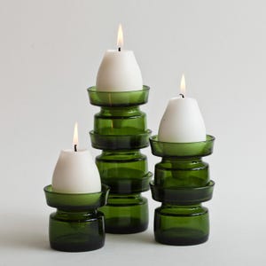 Può includere: Tre portacandele in vetro verde con candele bianche. I portacandele sono impilati in un design unico, con una base rotonda e una parte superiore svasata per tenere la candela. Le candele sono accese, con un bagliore soffuso.