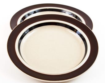 May include: Two round ceramic plates stacked on top of each other. Each plate features a light cream-colored center, a wide dark brown rim, and a thin black band separating the center from the outer edge. The plates have a smooth finish.