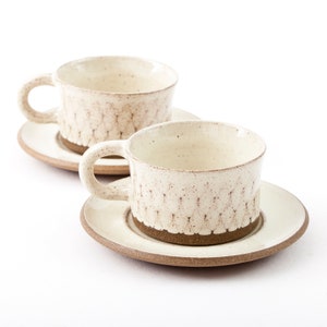 Puede incluir: Dos tazas de café de cerámica blanca con bordes marrones y textura moteada. Las tazas están sobre platillos a juego.