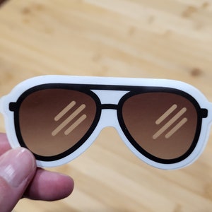 May include: A white sticker with a cartoon illustration of brown sunglasses with black frames.
