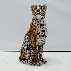 May include: A ceramic leopard figurine in a seated position. The spotted pattern features black spots on a white base, with orange and brown accents. The leopard has a detailed face and is a decorative item.