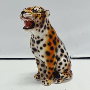 May include: A ceramic leopard figurine with an open mouth, showcasing its teeth. The spotted pattern features black spots on a cream and orange base. The figurine has green eyes and is posed in a seated position.