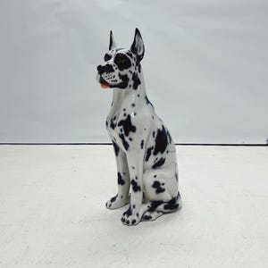 以下が含まれることがあります： 横を向いて座っているグレートデーンの犬のセラミック製置物。犬は黒と白で、黒い斑点があります。