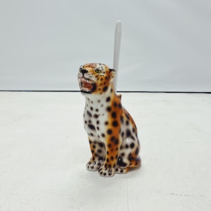 May include: A ceramic toilet brush holder shaped like a leopard with brown, black, and white spots. The leopard is sitting with its mouth open and has a white brush holder on its back.