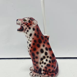 May include: A ceramic toilet brush holder shaped like a sitting leopard. The leopard has a red and black spotted pattern with green eyes and an open mouth. A white toilet brush extends from the back.
