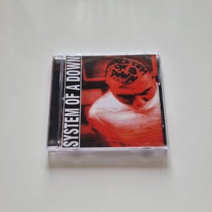 Könnte beinhalten: Eine CD-Hülle mit dem Albumcover von System of a Down. Das Cover ist überwiegend rot, mit dem Gesicht einer Person und dem Namen der Band auf dem Kopf. Der Text "SYSTEM OF A DOWN" ist vertikal auf der linken Seite des Covers aufgedruckt.