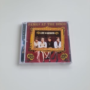 Puede incluir: Una caja de CD del álbum "Live in Denver" de Panic! at the Disco. La portada presenta una imagen enmarcada de los miembros de la banda. El fondo es rojo oscuro con detalles dorados e ilustraciones de rosas. La caja del CD es transparente.