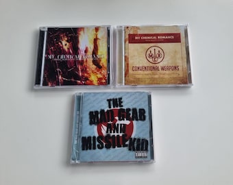 Peut inclure: Trois boîtiers de CD du groupe My Chemical Romance. Le boîtier supérieur gauche présente une image enflammée, le boîtier supérieur droit a un fond beige avec le logo du groupe et le boîtier inférieur a un motif de cible rouge.