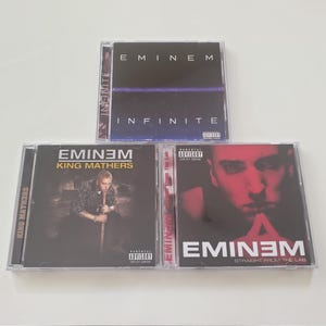 Puede incluir: Se muestran tres álbumes de CD de Eminem. El álbum superior se titula "Infinite" con un diseño azul oscuro y morado. El álbum de la izquierda es "King Mathers" con una foto de Eminem. El álbum de la derecha es "Straight From The Lab" con una imagen en tonos rojos de Eminem.