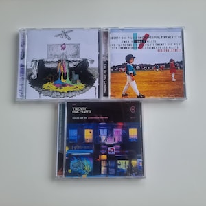 Può includere: Tre album CD dei Twenty One Pilots. Le copertine degli album presentano vari design, tra cui arte astratta, una scena di campo da baseball e un paesaggio urbano con luci al neon. I CD sono in custodie di plastica trasparente.